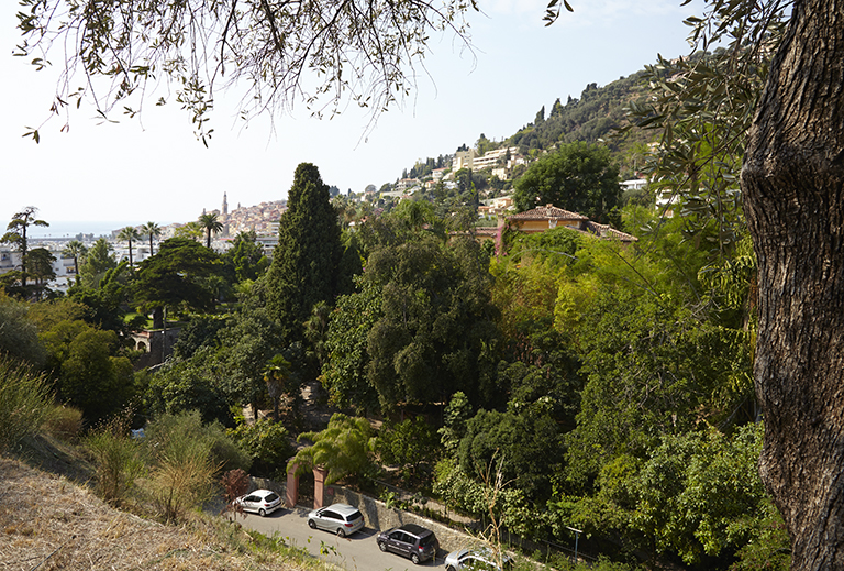 Vue d'ensemble du jardin prise depuis le Parc du Pian en direction du sud-ouest. Au premier plan, en contrebas, l'avenue Saint-Jacques et le portail de la propriété. Sur la droite, la maison. A gauche, le sentier de la Villa-Noël, le mur de soutènement à arcades et le jardin des plantes d'eau. A l'arrière-plan, la ville de Menton.