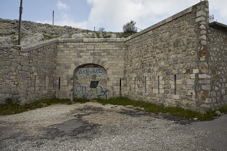 ancienne porte de l'ouvrage millésimée 1889 encadrée de flancs crénelés, vue extérieure