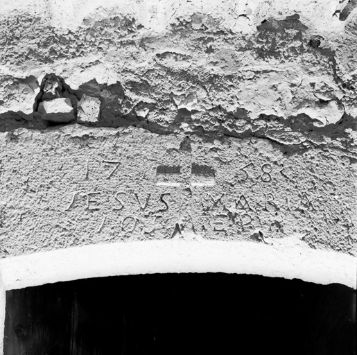 Elévation ouest, détail : la croix, la date et l'inscription portées sur le couvrement de la porte (logement), vue depuis l'ouest.
