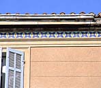 Détail du traitement décoratif de la façade : corniche en ciment, frise en carreaux de céramique, bandeau en ciment et encadrement de fenêtre en ciment moulé, enduit au fer.