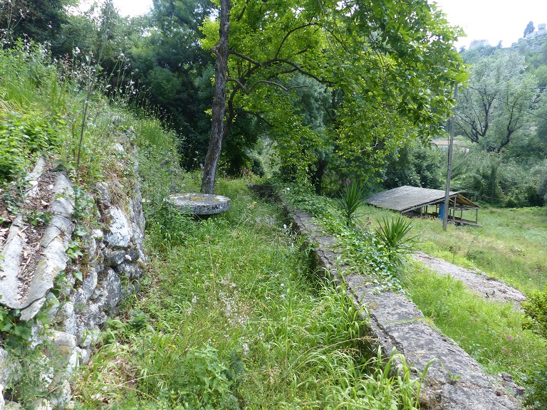 Vue d'ensemble prise du nord depuis la terrasse à l'arrière de la ferme (bâtiment 3). A droite, le mur de soutènement auquel est adossée la fontaine en-dessous ; au centre, une meule a été réutilisée en table de jardin ; à gauche, un mur en pierres sèches soutenant une terrasse de culture.