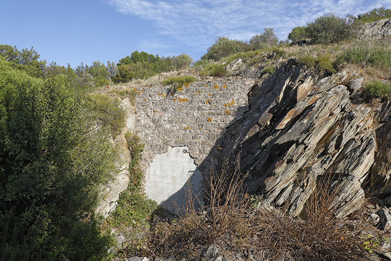 Mur de façade de l'entrée du magasin à poudres en caverne, porte arrachée et murée.