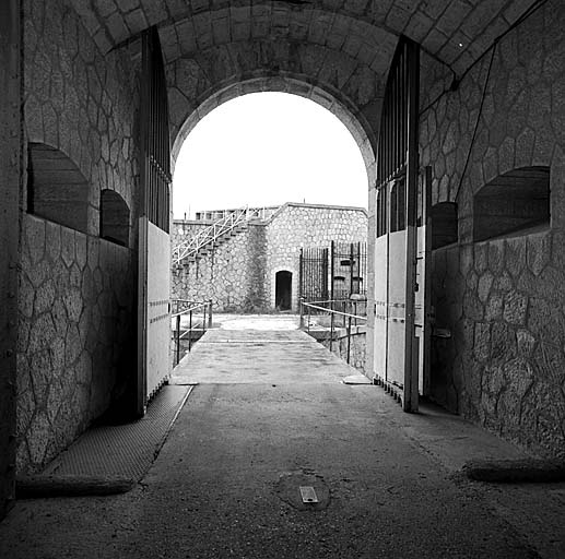 Entrée du fort. Le vestibule vu dans l'axe vers l'extérieur. Dans les parois, les créneaux de fusillade de défense intérieure, la porte-grille avec, à droite, son portillon. Au fond, ravelin et façade du bâtiment d (corps de garde avancé)