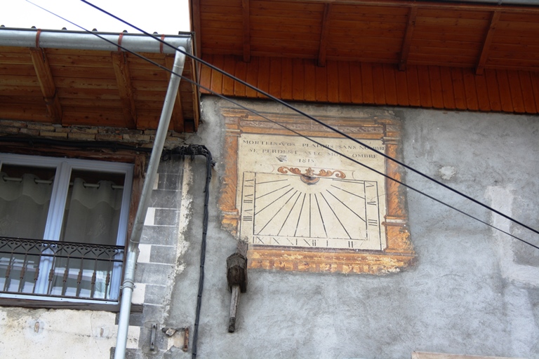 Le Village. Cadran solaire non restauré (daté 1815) avec devise, sur la façade d'une maison de la place principale (parcelle E 246) qui présente par ailleurs une ancre en bois. Remarquer la chaîne d'angle harpée peinte sur la façade mitoyenne (parcelle 245).
