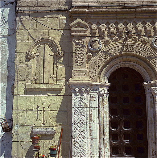 Détail de la façade de la chapelle, partie gauche. Décor de rocaille en trompe-l'oeil : faux appareil, portail roman, fausse fenêtre. Sous la fausse archère, date et signature de Stanislas Caillol.